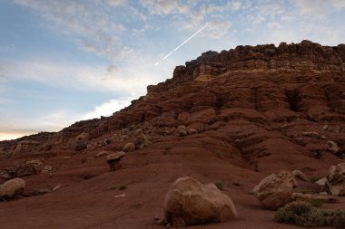 Canlı kırmızı kaya oluşumları açık bir gökyüzüne çarpıcı bir şekilde yükselir. Güneş batarken bir uçak görünür bir iz bırakır. Bu da jeolojik özellikleri aydınlatır..