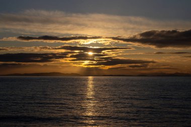 Çarpıcı bir günbatımı, sakin okyanus suları boyunca ılık altın bir ışık saçar ve gökyüzüne pastel bulutlar saçılır. Ufuk uzak kara oluşumlarını gözler önüne seriyor, barışçıl bir atmosfer yaratıyor..