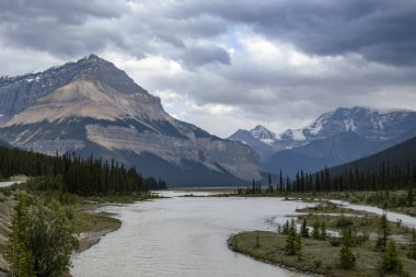 Sarmal bir nehir dramatik bulutlu bir gökyüzünün altında yükselen dağlarla çevrili güzel bir vadi boyunca akar. Doğal güzellik, bu sakin Kanada manzarasında sergileniyor..