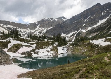 Yüksek irtifada sakin bir göl görkemli dağları ve çam ağaçlarını yansıtır. Kar lekeleri kalıyor ve temiz havada hafif bir esinti hissedebiliyorsun..
