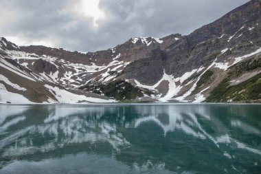 Dağlar görkemli bir şekilde sakin bir göl üzerinde, karla kaplı tepeler ve suları çevreleyen yemyeşil bir yer. Bulutlu gökyüzü huzurlu sahneye dramatik bir dokunuş ekler..