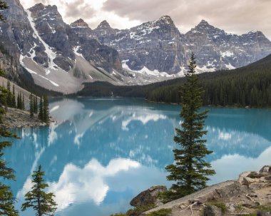Huzurlu Moraine Gölü, yemyeşil ormanlarla çevrili yüksek dağ tepelerini yansıtır. Sahne, yumuşak bulutlar ve canlı turkuaz sularla sabahın erken saatlerinin sükunetini yakalıyor..