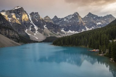Sakin bir göl, gün batımında çarpıcı dağ tepelerini ve gür ormanı yansıtır. Yumuşak ışık, bu doğal manzaranın huzur veren güzelliğini arttırır..
