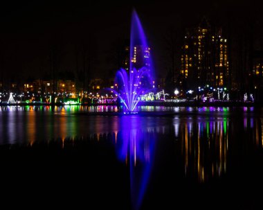 Çarpıcı bir çeşme, gece vakti bir parkta renkli ışınlar yayar. Etrafındaki ışıklar, suyun yüzeyine güzel yansıyan neşeli bir atmosfer yaratıyor..