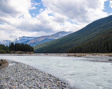 Berrak nehir, yüksek ağaçlar ve görkemli dağlarla çevrili kayalık bir kıyıdan akar. Huzurlu milli parktaki bulutlu bir gökyüzünün altında..