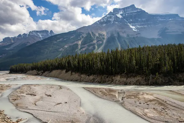 Çarpıcı bir manzarada, öğleden sonra erken saatlerde parçalı bulutlu bir gökyüzünün altında görünen görkemli dağlarla çevrili dolambaçlı bir nehir bulunur. Jasper Ulusal Parkı