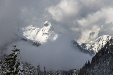 Görkemli kar örtülü dağ uzak bir dağlık bölgede dönen bulutların üzerinde yükselir. Evergreen ağaçları alttaki yamaçları kaplar, beyaz tepelerle çarpıcı bir kontrastlık yaratır..