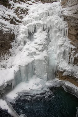 Göz kamaştırıcı bir şelalede buz oluşumları aşağıdaki karanlık bir su havuzuna dökülüyor. Karı çevreleyen karlar, el değmemiş kış manzarasını güzelleştirir, sakin bir atmosfer yaratır..