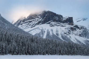 Sakin kış manzarası güneş ışığını yansıtan karla kaplı dağları yansıtırken, yüksek yeşil ağaçlar donmuş bir gölü çevreleyerek şafak vakti sakin bir atmosfer yaratıyor..