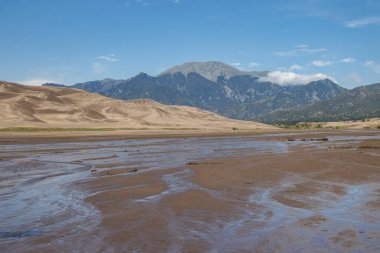 Bir nehir kumlu bir arazide esiyor ve uzak dağlar çarpıcı bir zemin oluşturuyor. Manzara, doğal güzellikleri yansıtan su ve toprağın canlı bir etkileşimine sahiptir..