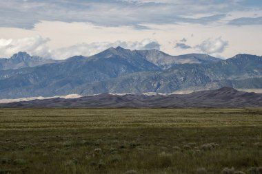Geniş çayırlar, üzerinde bulutların toplandığı görkemli bir dağ sırasına doğru uzanıyor. Manzara, öğleden sonra erken saatlerde manzaranın doğal güzelliğini yakalar..