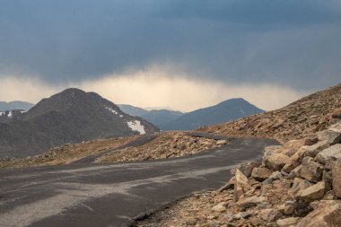 Engebeli arazide kayalık çıkıntılar ve uzak dağlarla çevrili dolambaçlı bir yol yılanı. Tepemizde kara bulutlar belirdi. Bölgedeki olası bir fırtınayı işaret ediyor..