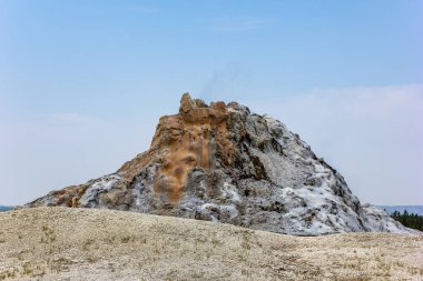 Canlı bir kaplıca buharla püskürüyor, kayalık yüzeyinde çarpıcı mineral yatakları sergiliyor. Etrafı seyrek bitki örtüsüyle çevrili açık mavi gökyüzü bu doğal harikanın güzelliğine katkı yapıyor..