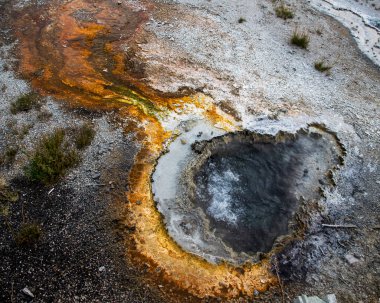 Parlak turuncu ve sarı mineral yatakları Yellowstone Ulusal Parkı 'nda kaynayan bir kaplıcayı çevrelemektedir. Canlı renkler koyu su ve kayalık araziyle tezat oluşturuyor..