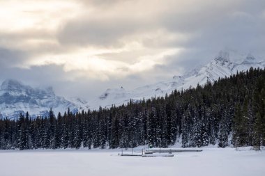 Sakin bir kış akşamında yoğun yeşil ağaçlarla çevrili karla kaplı bir göl sergilenir. Uzak dağlar bulutlarla örtülmüş, sakin bir atmosfer yaratıyor..