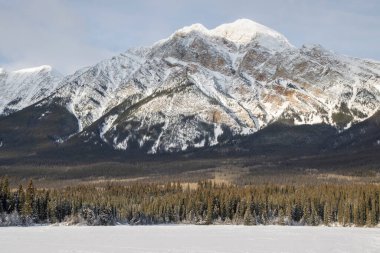 Karla kaplı zirveler, sık ve yeşil bir ormanın üzerinde donmuş bir gölün yanında, uzak bir kırsal alanda sakin bir kış manzarası sergiliyor..