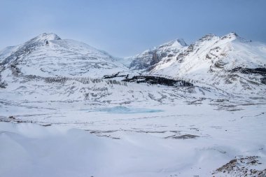 Karla kaplı görkemli dağlar berrak bir gökyüzüne karşı yükseliyor, karlı vadide görünen donmuş göl parçalarıyla, sakin bir kış atmosferi yaratıyor..
