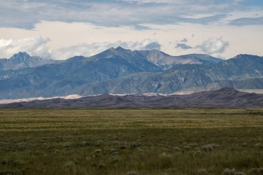Geniş çayırlar, üzerinde bulutların toplandığı görkemli bir dağ sırasına doğru uzanıyor. Manzara, öğleden sonra erken saatlerde manzaranın doğal güzelliğini yakalar..
