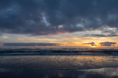 Güneş okyanusun üzerinde batarken, nemli kumu yansıtan ılık renkler gökyüzünü doldurur. Yumuşak dalgalar kıyıya vurur ve bir yaz akşamında huzurlu bir atmosfer yaratır..