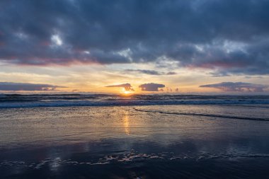 Gün batımının nefes kesici görüntüsü okyanusu altın ışıkla yıkayan dalgalar yavaşça kıyıya vuruyor. Gökyüzü dramatik bulutlar tarafından kenarlı sıcak renkler sergiliyor.