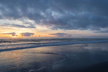 Güneş alacakaranlıkta uzak bulutların ardında batar ve suyun yüzeyine sıcak bir ışıltı yayar. Dalgalar yavaşça sahile çarpıyor ve bu sakin sahil bölgesinde huzurlu bir ortam yaratıyor..