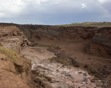 Kuru bir nehir yatağını çevreleyen ve kurak araziyi aydınlatan nefes kesici bir manzara sergiliyor. Tepemizde kara bulutlar beliriyor, muhtemel yağmuru işaret ediyor..