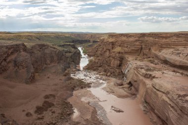 Göz kamaştırıcı bir kanyon, bulutlarla dolu bir gökyüzünün altındaki kayalık uçurumlarda kıvrılan kuru bir nehir yatağını gözler önüne seriyor. Engebeli arazi, önceki su akışını işaret eden toprak tonlarını ve seyrek bitki örtüsünü sergiliyor..