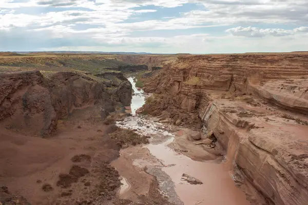 Göz kamaştırıcı bir kanyon, bulutlarla dolu bir gökyüzünün altındaki kayalık uçurumlarda kıvrılan kuru bir nehir yatağını gözler önüne seriyor. Engebeli arazi, önceki su akışını işaret eden toprak tonlarını ve seyrek bitki örtüsünü sergiliyor..
