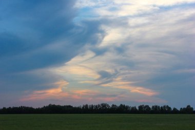 Nefes kesici bir manzara, mavi, pembe ve beyaz tonlarında dönen bulutları sergiliyor, sakin yeşil bir tarlanın üzerinde ve gün batımında karanlık bir orman siluetinde..