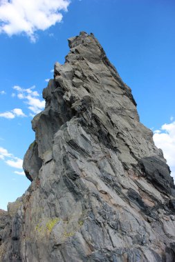 Büyük kaya oluşumu parlak mavi bir gökyüzüne karşı belirgin bir şekilde duruyor, engebeli dokular ve gölgeler sergiliyor. Kıyı manzarası manzaranın doğal güzelliğini arttırır..