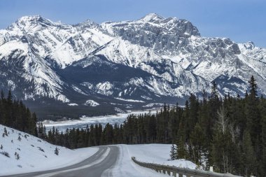 Kıvrımlı bir yol, yüksek dağlarla çevrili karlı bir araziyi kesiyor. Çam ağaçları yol kenarında, ve gökyüzü açık, bu da sakin bir kış günü olduğunu gösterir..