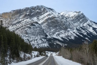 Bir araba uzak bir dağ bölgesinden geçerken kar, virajlı bir yol boyunca zemini kaplar. Yüksek tepeler berrak bir gökyüzünün altında çarpıcı bir zemin oluşturur..