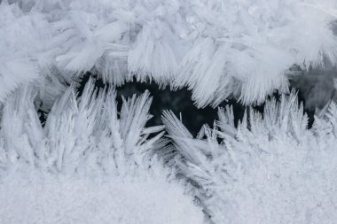 Narin buz kristalleri karanlık bir yüzeyi kaplayarak kışın doğanın sanatını sergiliyor. Karmaşık tasarımlar mevsimin soğuk atmosferini ve güzelliğini yansıtıyor..
