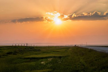 Güneş sessiz bir otoyol boyunca batarken altın ışık ufuk boyunca yayılıyor. Bereketli yeşil alanlar sahneyi çevreleyerek, akşamın huzurlu atmosferini güçlendiriyor..