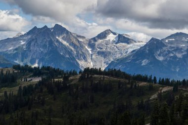 Uzun dağlar, dağınık bulutlardan oluşan bir zemin üzerinde heybetli bir şekilde yükselirken, parlak yeşil bir manzara ön planı doldurur. Sahne doğanın güzelliğini ve sükunetini sergiliyor..