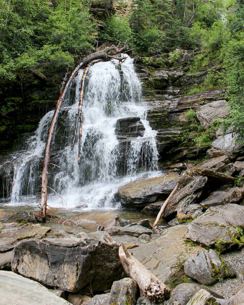 A stunning waterfall tumbles over large rocks, surrounded by vibrant greenery. The sun shines down, creating a serene atmosphere in a peaceful forest setting.