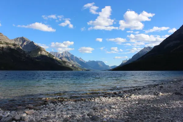 Sakin bir dağ gölü, tüylü bulutlarla benekli parlak mavi bir gökyüzünün altındaki çarpıcı manzarayı yansıtır. Kayalık kıyı şeridi sakin sulara götürür, keşifleri davet eder..