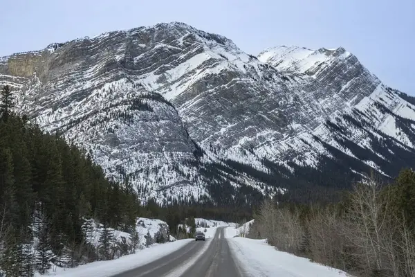 Bir araba uzak bir dağ bölgesinden geçerken kar, virajlı bir yol boyunca zemini kaplar. Yüksek tepeler berrak bir gökyüzünün altında çarpıcı bir zemin oluşturur..