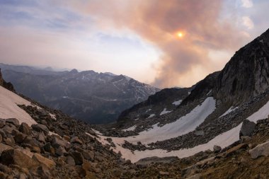 Nefes kesici bir manzara, güneş batarken dumanlı bir gökyüzünün altında kar ve engebeli kayalarla dolu bir vadiyi gösteriyor. Uzakta dağlar belirir, sakin ama kasvetli atmosferi güçlendirir..