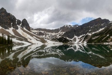 Sakin bir dağlık göl, engebeli arazi ve kar parçalarıyla çevrili görkemli dağ tepelerini ve bulutlu gökyüzünü yansıtır. Doğanın güzelliği bu huzurlu ortamda büyülenir..
