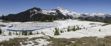Arka planda görkemli zirveler yükselirken, buzul gölü güneş ışığını yansıtıyor..