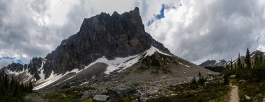 Bulutlu bir gökyüzüne karşı yükselen yüksek dağlar engebeli araziler ve kar yığınları sergiliyor. Manzarada kayalık yamaçlar ve canlı yeşillikler arasında dolambaçlı bir yol bulunur..