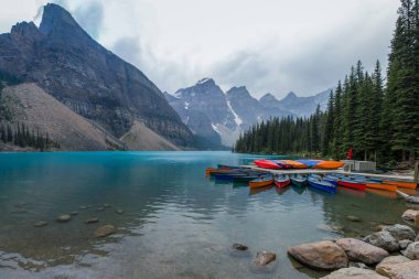 Renkli kanolar sakin bir göl kenarındaki rıhtımda, bulutlu bir gökyüzünün altında arka planda yükselen dağlarla düzgünce düzenlenir. Huzurlu sahne doğanın güzelliğini sergiliyor..
