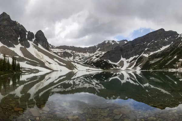 Sakin bir dağlık göl, engebeli arazi ve kar parçalarıyla çevrili görkemli dağ tepelerini ve bulutlu gökyüzünü yansıtır. Doğanın güzelliği bu huzurlu ortamda büyülenir..