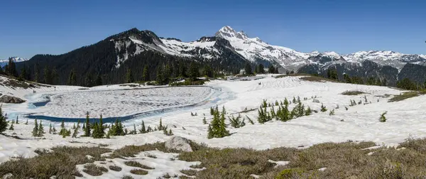 Arka planda görkemli zirveler yükselirken, buzul gölü güneş ışığını yansıtıyor..