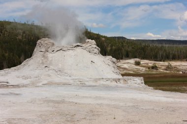 Bir gayzer patladı ve Yellowstone Ulusal Parkı 'nda havaya buhar püskürdü. Engebeli arazi yumuşak toprak tonlarını ve berrak mavi gökyüzünün altında uzak dağları barındırıyor, doğanın harikalarını vurguluyor..