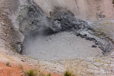 Jeotermal bir bölgede fokurdayan çamur havuzu jeotermal aktivite tarafından yüzeye itilen gri, kalın çamuru içerir. Çevresindeki manzara, berrak bir gökyüzünün altında erozyon ve mineral yatakları sergiliyor..