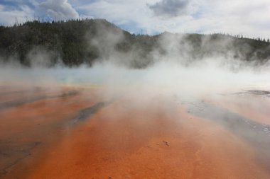 Parlak turuncu renkler Yellowstone Ulusal Parkı 'ndaki kaplıcalardan yükselen buharla karışır. Yeşil dağlarla çevrili bu doğa harikası jeotermal aktivitenin özünü yakalar..