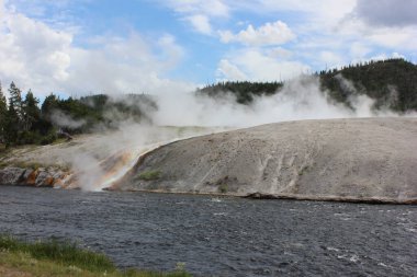 Güneşli bir yaz öğleden sonrasında Yellowstone Ulusal Parkı 'ndaki bir nehir kıyısı boyunca renkli jeotermal oluşumlardan gelen buhar dalgaları bölgenin eşsiz jeolojik aktivitesini ve doğal güzelliğini gözler önüne seriyor..