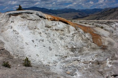 Canlı turuncu ve beyaz mineral yatakları mavi gökyüzünün altındaki Yellowstone Ulusal Parkı 'nın engin manzarasında kayalık bir oluşumda çarpıcı desenler oluşturur..